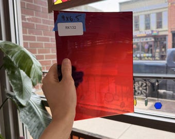 Puede incluir: Una lámina de vidrio rojo transparente, de 22,86 x 16,51 cm, se sostiene a la luz. La etiqueta del vidrio dice "RK132". El vidrio está frente a una ventana con un vitral.