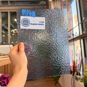 May include: A textured, clear glass sheet labeled "English Muffle" with dimensions of 11 x 8.5 inches. The glass has a bubbly, irregular surface. A blue tag indicates "G47". The sheet is held up in front of a window.