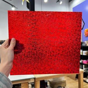May include: A vibrant red, textured sheet of material. The sheet has a reflective, patterned surface. The material is being held up in front of a neutral background. The sheet is rectangular in shape.
