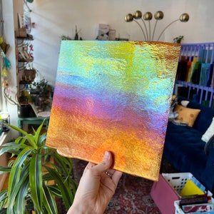 Puede incluir: Una pieza cuadrada de vidrio iridiscente con una superficie texturizada. El vidrio tiene un degradado de colores, comenzando con un amarillo claro en la parte superior y pasando a naranja, rojo y morado en la parte inferior.