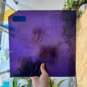 May include: A square sheet of textured, deep purple stained glass. A blue label with white text is affixed to the top left corner. The glass is held by a hand, with plants and a window visible in the background.