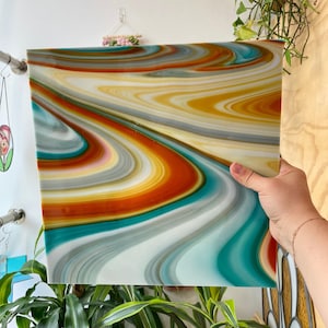 Könnte beinhalten: Ein quadratisches, abstraktes Kunstwerk mit einem wirbelnden Farbmuster. Das Design zeigt Bänder in Orange, Gelb, Türkis, Grau und Weiß, die einen fließenden, marmorierten Effekt erzeugen. Das Kunstwerk wird von einer Hand gehalten.