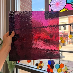May include: A square sheet of translucent stained glass in a deep magenta and purple hue. The glass features a textured, rippled surface that catches the light, creating variations in color intensity across the pane's flat, uniform surface.