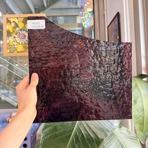 May include: A rectangular sheet of dark purple stained glass with a textured, rippled surface. A white label in the top left corner displays the text RL922 and 12x10.5 inches. The glass is translucent, showing a deep violet hue when light passes through.