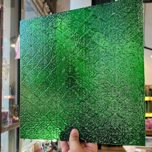 Könnte beinhalten: Ein quadratisches, smaragdgrünes Glaspaneel mit einem strukturierten, sich wiederholenden floralen Muster. Das Glas wird hochgehalten, wodurch seine Transparenz und das komplizierte Design zur Geltung kommen. Das Paneel ist wahrscheinlich für dekorative oder architektonische Zwecke bestimmt.