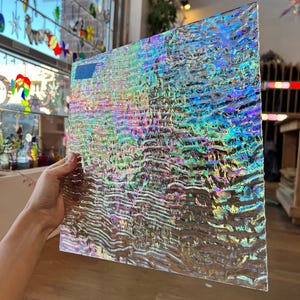 May include: A rectangular sheet of iridescent glass with a textured finish. The glass shimmers with a range of colours, including blues, greens, and purples. The glass is held, highlighting its size and the light reflecting across its surface.