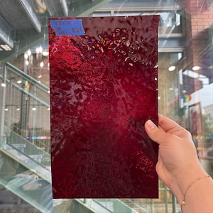 以下が含まれることがあります： テクスチャ加工されたルビーレッドガラスの長方形のシート。ガラスは波状の表面と光沢のある仕上がりです。左上隅にある青い付箋には「11.5 x 7」と他のテキストが書かれています。ガラスは手で持たれています。