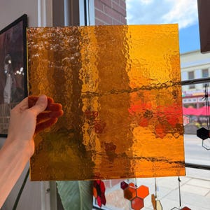 Puede incluir: Una hoja cuadrada de vidrio tintado translúcido de color ámbar con una superficie texturizada y ondulada. El vidrio se sostiene frente a una ventana, permitiendo que la luz natural pase a través y resalte el patrón irregular y ondulado del material.