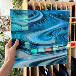 Könnte beinhalten: Ein quadratisches Glasstück mit einem wirbelnden Muster aus Blau-, Weiß- und Grüntönen. Das Glas wird von einer Hand gehalten, wodurch die lebendigen Farben und das fließende Design zur Geltung kommen. Es scheint sich um ein dekoratives Kunstwerk zu handeln.