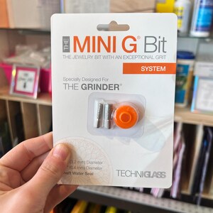 May include: A package for a "MINI G Bit" jewelry tool, designed for "THE GRINDER". The package is white with orange and gray accents, and includes the tool itself, which has an orange component and two silver cylindrical pieces. The text "TECHNIGLASS" is printed on the package.
