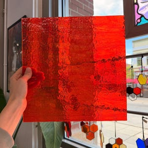 Könnte beinhalten: Ein quadratisches Blatt aus leuchtend rotem Buntglas mit einer texturierten, gewellten Oberfläche. Das Glas ist durchscheinend und lässt Licht hindurch, wodurch Variationen in der tiefroten Farbe und das unebene, organische Muster sichtbar werden.