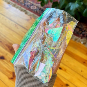 May include: A clear plastic bag filled with iridescent holographic paper scraps in various shapes and sizes. The bag is partially open and the paper scraps are visible.