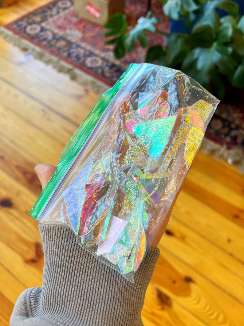 May include: A clear plastic bag filled with iridescent holographic paper scraps in various shapes and sizes. The bag is partially open and the paper scraps are visible.
