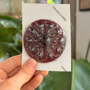 Può includere: Un oggetto decorativo rotondo, rosso scuro, con una superficie strutturata e un punto centrale scuro. Montato su una carta bianca con la scritta "Made in Germany".