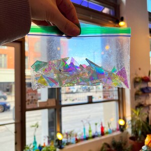 May include: A clear plastic bag filled with iridescent, triangular glass pieces. The glass pieces are various shades of blue, green, and purple.