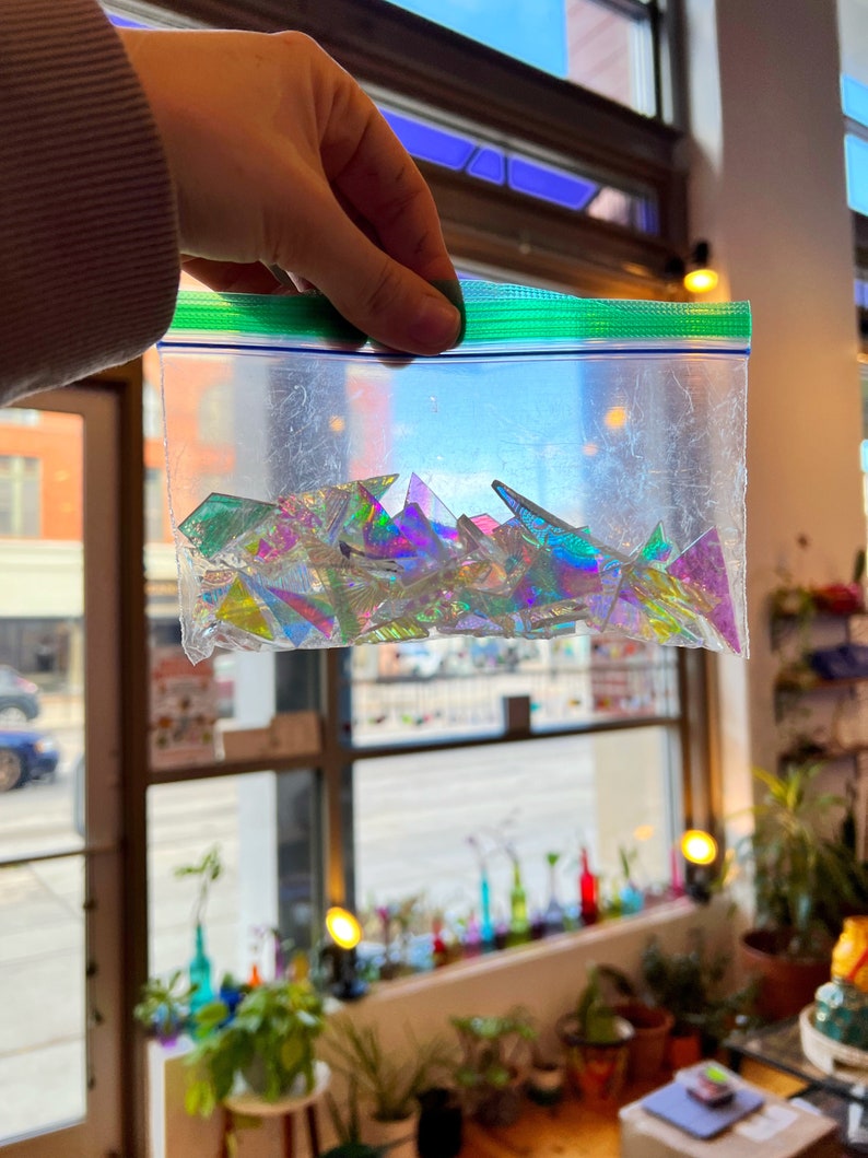 May include: A clear plastic bag filled with iridescent, triangular glass pieces. The glass pieces are various shades of blue, green, and purple.