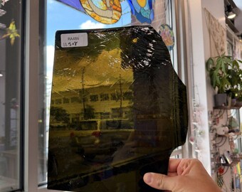 May include: A rectangular sheet of dark olive green stained glass with a textured, rippled surface. A white rectangular sticker in the top left corner displays the text RA486 and 11.5x8. The glass is semi-transparent, showing reflections of the outdoors.