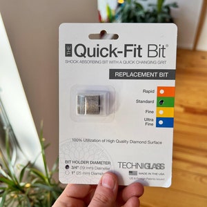 May include: A white packaging for a Quick-Fit Bit replacement bit for a diamond drill. The packaging shows a silver bit, the text "Quick-Fit Bit", and the text "Shock Absorbing Bit with a Quick Changing Grit". The packaging also shows the text "Replacement Bit" and lists different grit options: Rapid, Standard, Fine, Ultra Fine. The packaging also shows the text "100% Utilization of High Quality Diamond Surface" and lists the bit holder diameter options: 3/4" (19 mm) Diameter and 1" (25 mm) Diameter. The packaging also shows the text "Techniglass" and "Made in the USA".