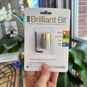 May include: A package for "The Brilliant Bit" features a gold and silver diamond grit bit with a black hex key. The text reads "THE GOLD STANDARD BIT WITH A DIAMOND GRIT" and "FITS ALL GRINDERS 3/4" (19 mm) Diameter 1" (25 mm) Diameter".