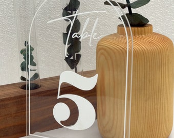May include: Clear acrylic table number holder with a white number 5 and the word "Table" written in cursive. The holder is on a wooden surface with a vase of greenery.