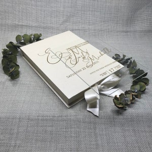 Puede incluir: Una caja de acr&iacute;lico transparente con una cinta de sat&eacute;n blanca y una tarjeta blanca en su interior. La tarjeta tiene una inscripci&oacute;n dorada que dice "Together with their families and friends, Emily & Michael, Saturday 22 May 2021, Invite you to their wedding celebration, 4:00 pm, Saturday, 17, 2021".