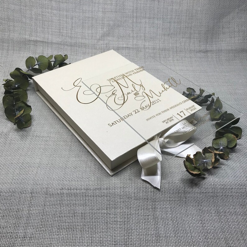 Puede incluir: Una caja de acr&iacute;lico transparente con una cinta de sat&eacute;n blanca y una tarjeta blanca en su interior. La tarjeta tiene una inscripci&oacute;n dorada que dice "Together with their families and friends, Emily & Michael, Saturday 22 May 2021, Invite you to their wedding celebration, 4:00 pm, Saturday, 17, 2021".