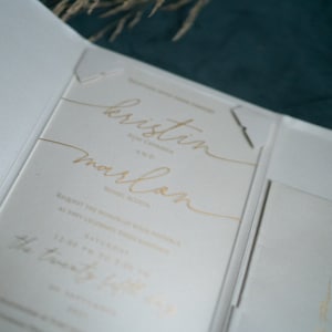 May include: A white wedding invitation with gold lettering. The invitation reads "kristin and marlon" with the date and time of the wedding below.