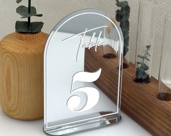 May include: Silver mirror table number with a white arch and the number 5 in a script font. The text "Table" is written in a script font above the number.
