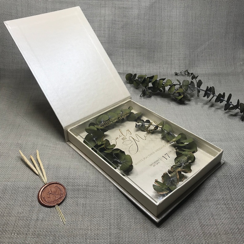 Puede incluir: Una caja blanca con un inserto de acr&iacute;lico transparente que contiene una invitaci&oacute;n de boda con vegetaci&oacute;n seca y el texto "Emily and Matt" en letras doradas. La invitaci&oacute;n dice "Invite you to their wedding" e incluye la fecha "September 17".