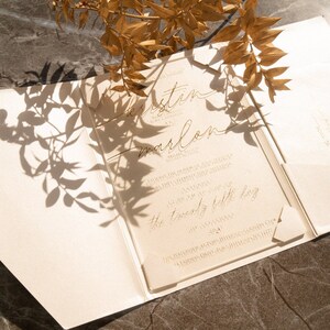 May include: A clear acrylic wedding invitation with gold lettering. The invitation reads "Kristin and Marlon" and includes the date and time of the wedding. The invitation is sitting on a white background.
