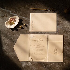 May include: A beige wedding invitation with a clear acrylic insert featuring the names "kristin" and "marlon" in gold script. The invitation includes details about the wedding ceremony and reception, including the date, time, and location. The invitation is folded and placed on a brown surface.