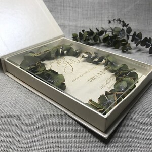 Puede incluir: Una caja de acr&iacute;lico transparente con un interior blanco, que contiene una invitaci&oacute;n de boda con letras doradas y vegetaci&oacute;n seca. La invitaci&oacute;n dice "Invite you to their wedding" e incluye la fecha "Saturday, 17th of April".