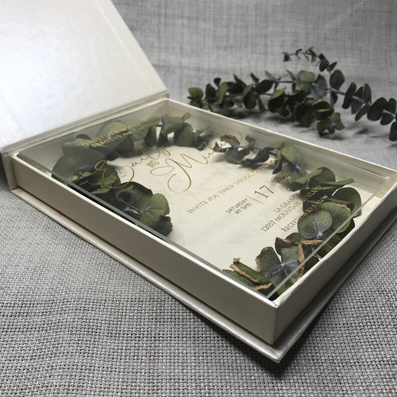 Puede incluir: Una caja de acr&iacute;lico transparente con un interior blanco, que contiene una invitaci&oacute;n de boda con letras doradas y vegetaci&oacute;n seca. La invitaci&oacute;n dice "Invite you to their wedding" e incluye la fecha "Saturday, 17th of April".