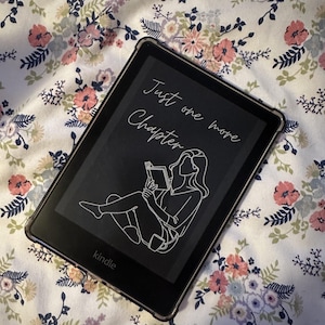Può includere: Un e-reader Kindle nero con una custodia protettiva trasparente, che mostra il testo "Just one more Chapter" e un disegno a tratto di una persona che legge. L'e-reader è appoggiato su un tessuto a fantasia floreale con fiori rosa, pesca e blu.