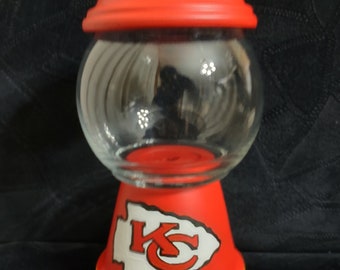 Op de afbeelding: Een rode, witte en gele keramische snoepvaas met een glazen bol en een logo van de Kansas City Chiefs op de voet.