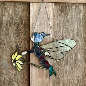 May include: A stained glass fairy with blue wings and a blue hat holding a yellow flower. The fairy is hanging from a chain on a wooden background.