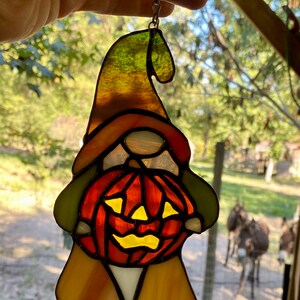 May include: A stained glass gnome holding a jack-o'-lantern. The gnome is wearing a green hat, orange pants, and a white shirt. The jack-o'-lantern is orange with a yellow face.