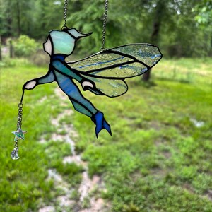 May include: A stained glass fairy with blue and green wings, holding a star and moon charm. The fairy is hanging from a silver chain against a blurred green background.