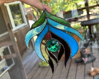 May include: A stained glass peacock feather suncatcher with a green jewel in the center. The feather is made of blue, green, and red glass.