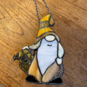 May include: A stained glass gnome ornament with a yellow hat, brown beard, and a white face. The gnome is holding a brown and gold basket in its left hand. The ornament is hanging from a silver chain.
