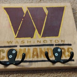 May include: Wooden wall hook with two black metal hooks. The hook is painted with a burgundy "W" and the words "WASHINGTON COMMANDERS" in gold.