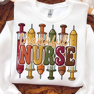Op de afbeelding: Een wit sweatshirt met lange mouwen en een grafische print van zes verticale injectiespuiten in verschillende kleuren, waaronder rood, geel, oranje, groen en bruin. De tekst "thankful NURSE" is in een gestileerd, meerkleurig lettertype over de spuiten gedrukt.
