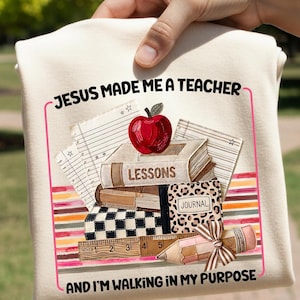 Könnte beinhalten: Ein cremefarbenes T-Shirt mit einer Grafik aus gestapelten Büchern, einem roten Apfel, einem Lineal und einem Bleistift. Der Text "JESUS MADE ME A TEACHER" und "AND I'M WALKING IN MY PURPOSE" steht in schwarzer Schrift, umrahmt von einem rosa Rand.