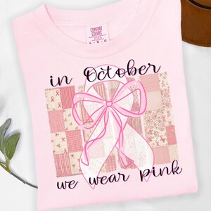 以下が含まれることがあります： 「in October we wear pink」の文字が入った薄いピンク色のTシャツ。ピンクのリボンとパッチワークの背景が特徴です。Comfort Colors製です。