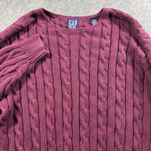 May include: A deep maroon long-sleeve jumper featuring a vertical cable knit pattern throughout the garment. The jumper has a round neckline and a blue rectangular tag at the inner collar with the white text GAP printed in capital letters.