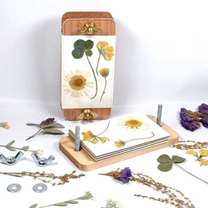 May include: A wooden flower press with a set of four pressed flowers. The press is made of wood and has a metal screw to tighten the press. The flowers are pressed between two pieces of cardboard. The flowers are a variety of colors, including yellow, white, and green.