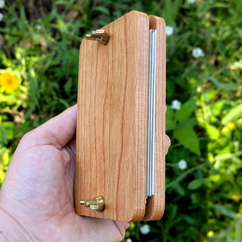 May include: A wooden book press with brass hardware. The press is made of light brown wood and has a rectangular shape. The metal bars are visible on the side. The press is held in a hand against a background of green foliage.