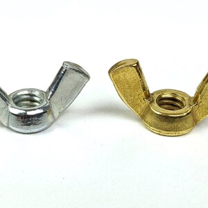 May include: Two wing nuts, one silver and one gold, are displayed against a white background. The wing nuts have a threaded center and two flat, wing-shaped extensions for easy tightening by hand. These fasteners are commonly used in various applications.