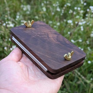 May include: A dark brown wooden plant press with brass wing nuts. The press has a rectangular shape with a clear plastic strip along one edge. The wood grain is visible. The press is designed for pressing flowers and plants.