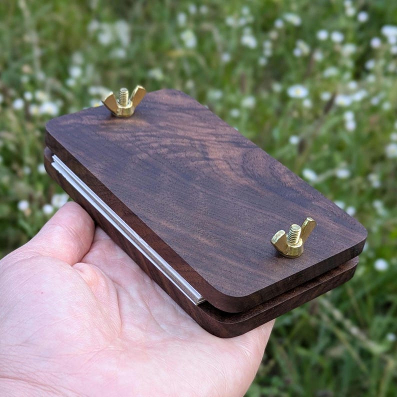 May include: A dark brown wooden plant press with brass wing nuts. The press has a rectangular shape with a clear plastic strip along one edge. The wood grain is visible. The press is designed for pressing flowers and plants.